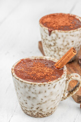 Hot chocolate with cocoa powder and cinnamon stick in ceramic mug on light background with copy space