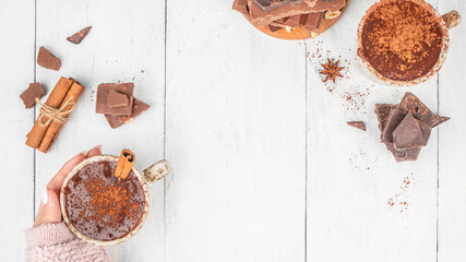 Top view of hot chocolate with cocoa powder and cinnamon held in hand on white wooden background with copy space
