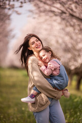 Fototapeta premium Mother and daughter share a warm hug and bright smiles in a sunlit almond orchard in bloom, enjoying springtime bonding among petals, trees and green grass