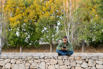 A pensive young man dressed in a green hoodie and jeans sits on a white stone wall during golden...