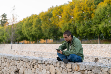 A contemplative brunette man in a green hoodie and jeans sits cross-legged on a white stone wall at dusk, his silhouette sharply defined against the fading light as he intently reads an ebook.  © Araguatai