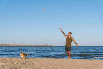 Man with tattoos playing fetch with his dog on a sunny beach, throwing a ball high into the air