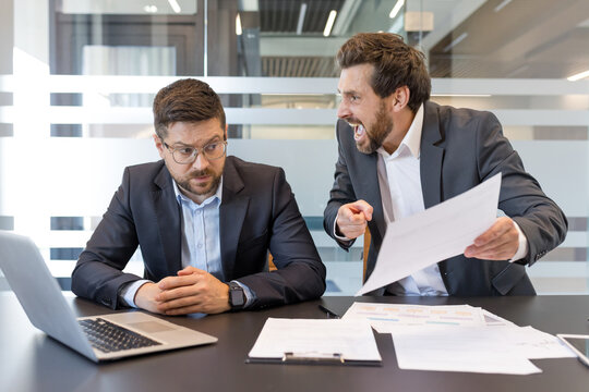 Male executive aggressively berating an overwhelmed subordinate at a cluttered desk, capturing workplace conflict, stress, bullying, and a tense, toxic office confrontation