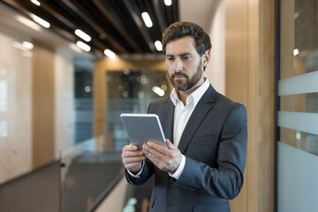 Fototapeta premium Confident businessman in a modern office hallway using a digital tablet to review data and manage tasks, illustrating technology-driven productivity and professional leadership