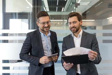 Two smiling professional businessmen standing in a modern office, collaborating and reviewing documents on a clipboard while one holds a tablet, focusing on partnership and planning