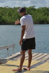 Fototapeta premium man walking on the pier, man in white t-shirt and black shorts standing on wooden pier, person gesturing near calm river water in tropical nature, man wearing baseball cap looking at lagoon, back view