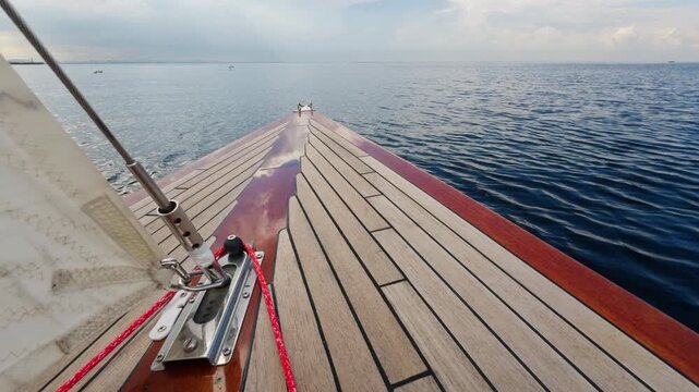 the bow of a beautiful wooden sailboat sailing through calm water, the glossy red side of the boat, the chrome details of the boat's equipment, the teak deck