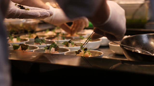 Professional chef in gloves using precision tweezers to garnish gourmet appetizers in a commercial kitchen. High-end food styling, culinary production, and technical plating for fine dining.