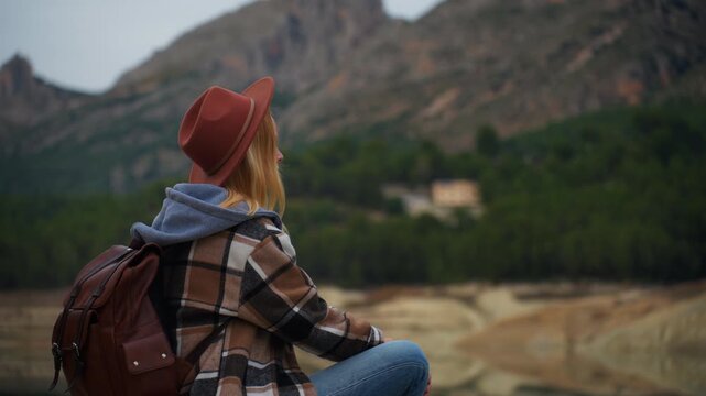 Young woman with hat and backpack sit by lake, enjoy tranquility and beauty of mountain scenery during trip. Adventurous journey and connection with nature