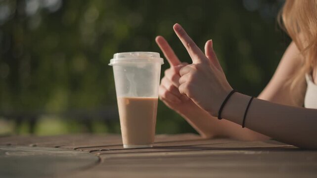 Young white woman taps iced coffee cup on wooden table, fingers counting rhythm like street dancer, sunlight filtering through park trees, shallow bokeh background, warm summer tones, casual bracelet