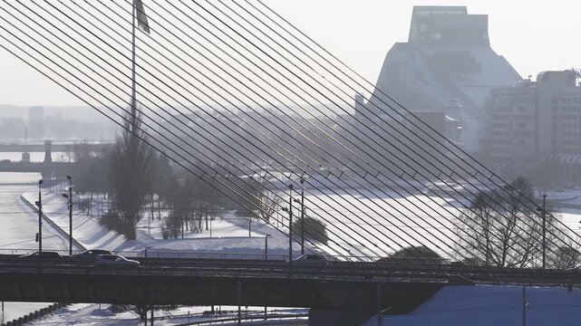 Aerial over the frozen Daugava in Riga shows Vansu Bridge with diagonal cables, Latvian National Library, cars and a van crossing, snow embankments, long winter light.