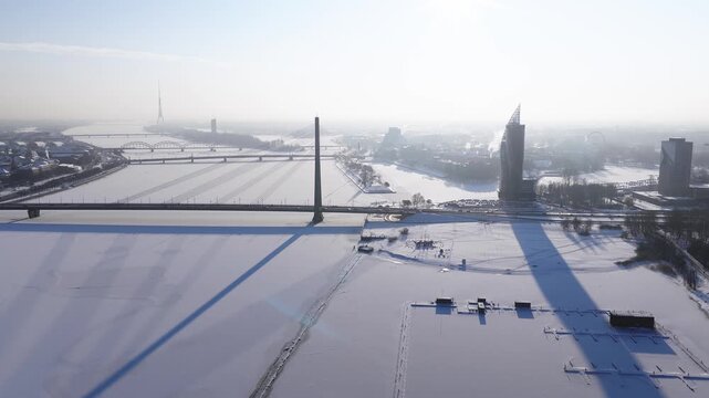 Aerial view in Riga, Latvia shows Vansu Bridge centered on the Daugava, Riga TV Tower on a misty horizon, Old Town spires, modern high rises, and frozen docks under snow.
