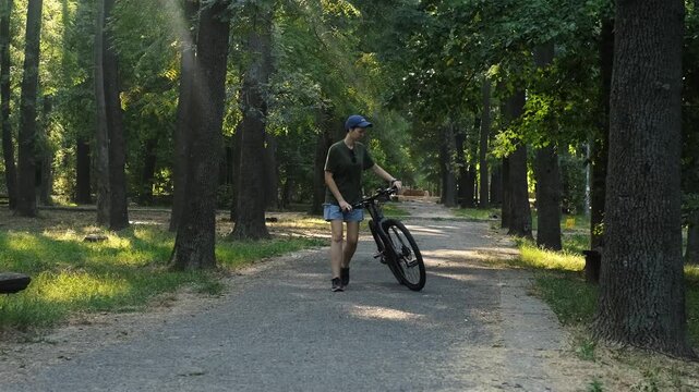 A woman walks with a bicycle through a shaded forest park path on a summer day. The moment conveys relaxation, motion and a natural outdoor routine.
