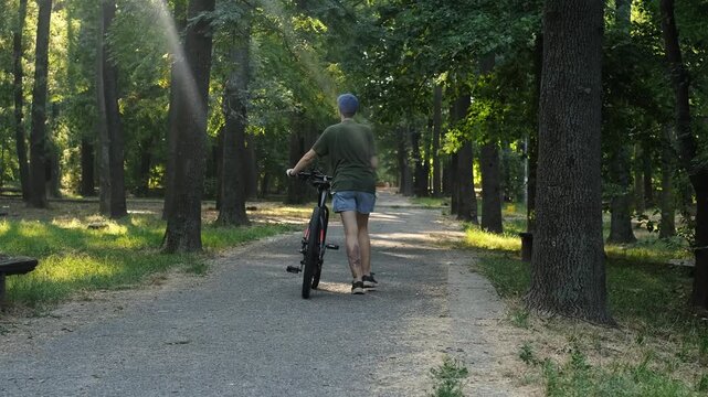 A woman walks with a bicycle through a shaded forest park path on a summer day. The moment conveys relaxation, motion and a natural outdoor routine. Rear view
