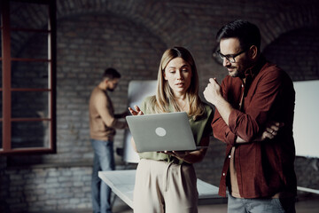 Two young entrepreneurs in casual clothes looking at a laptop screen and discussing a project, while a colleague works in the background of a modern creative office.