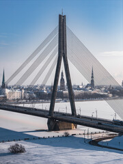 Obraz premium Aerial view of Vansu Bridge over the frozen Daugava River in Riga, Latvia. The A shaped pylon and cables frame Riga Cathedral and St. Peter's spire in clear cold light.