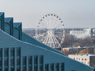 A Ferris wheel rises above treetops and city blocks, framed by the National Library of Latvia in...