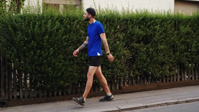A man in sportswear walks steadily along a calm residential street beside a dense green hedge, maintaining a relaxed but focused pace. The scene highlights daily physical activity and urban lifestyle