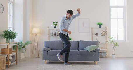 Happy man dancing to cheerful music funny goofy dance at home. Crazy excited guy in casual sneakers, jeans and blue shirt singing during weird silly movements with playful energy in living room