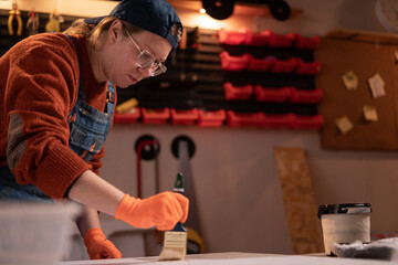 Woman in workwear and glasses painting object with brush in home garage with tools hanging on wall....