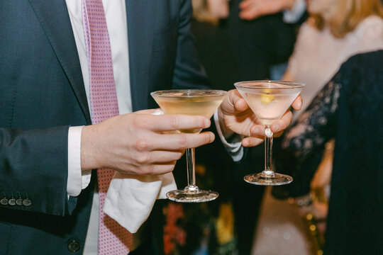 Two martini cocktails held by a man in a suit at a social gathering in an indoor venue during evening hours