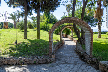 Stone archway over a pedestrian path in a lush green city park. Scenic garden landscape with paved walkway, grass lawn, and tropical trees. September, Antalya, Turkey.