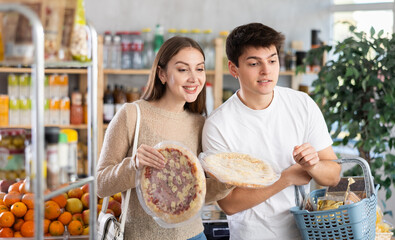 Happy young wife and husband choose large and fresh pizza in big supermarket in department of...