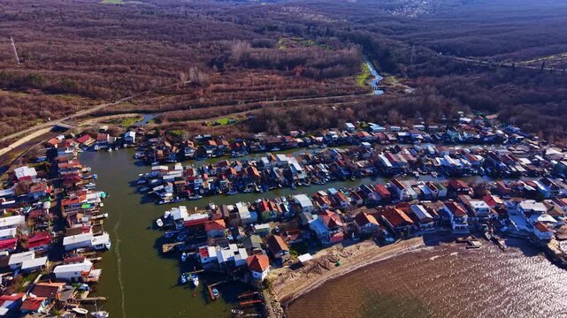 Houses sit along the water in a village, with boats docked nearby. Trees line the area, and the scene shows natural surroundings, creating a view from above.