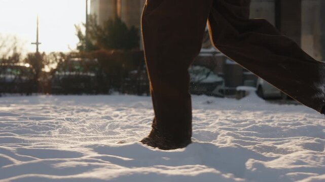 Close-up gimbal follow shot of man walking through fresh snow in winter city park. Side view brown pants and black leather boots step through deep snow on cold sunny day. Snowy weather on city street