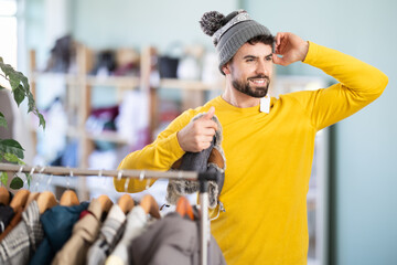 Male customer is trying on winter hat during shopping in store, examining reflection in mirror. Fitting room, stock clothing store.