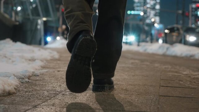 Rear view gimbal follow shot of man walking alone across city street sidewalk on winter evening. Male feet steps close-up, brown pants leather boots. Urban lifestyle, snowy weather in modern city
