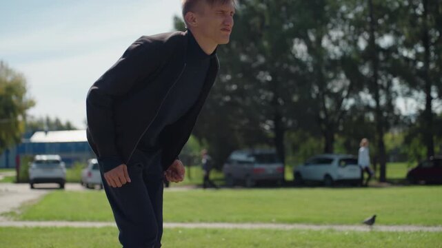 young man bent forward in park, casual jacket, focused gaze toward ground near pigeon, intermittent pause as if catching breath, playful head tilt, suburban background with parked cars and trees, soft