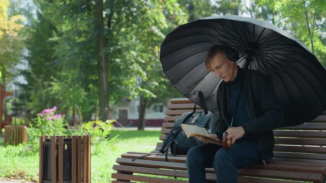 caucasian actor rehearsing script under umbrella, animated facial expressions and expressive hand gestures while reading lines, black umbrella used as prop, backpack with costume pieces at side,