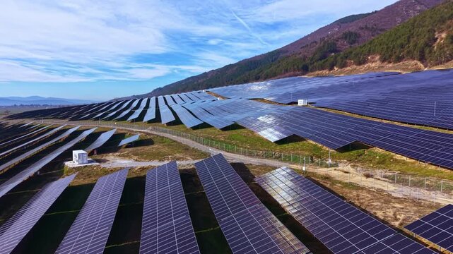 Large field of solar panels stretches across a sloped land with hills in the background under a bright sky. Panels capture sunlight for energy production.