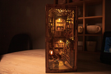 Magical wooden book nook miniature library diorama glowing with warm lights in the dark. Intricate bookshelf alleyway with tiny books, ladder, and vintage decor. Enchanting handmade room box model. © Alisa