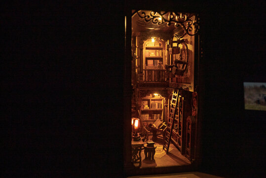 Magical wooden book nook miniature library diorama glowing with warm lights in the dark. Intricate bookshelf alleyway with tiny books, ladder, and vintage decor. Enchanting handmade room box model.