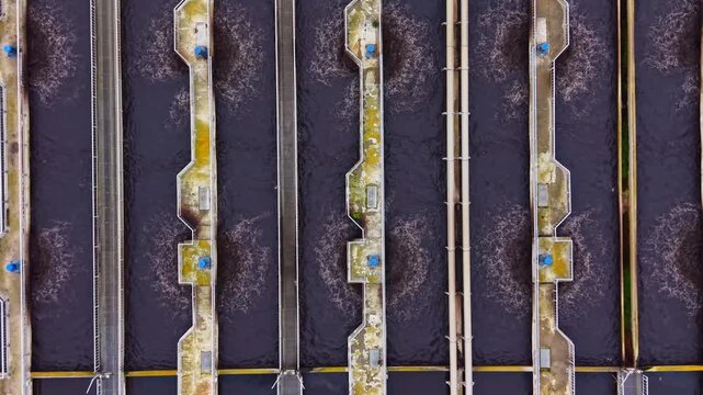 Water treatment facility shows several large, rectangular tanks filled with dark water, with water flowing in channels between them during the day.