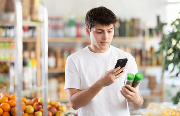 Young guy using smartphone to scan and and compare two grounded herbal spices in supermarket aisle, comparing prices, ingredients, and online reviews to choose best option for gastronomic treat