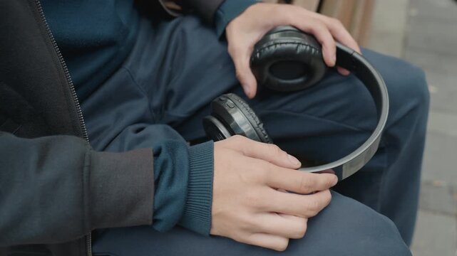 hands holding headphones on lap, commuter pauses on city bench adjusting earcup and leather cushion, denim trousers and textured sweater sleeve visible frames alternate between street musician