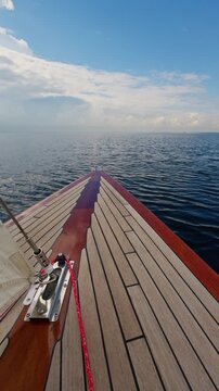 the bow of a beautiful wooden sailboat sailing through calm water, the glossy red side of the boat, the chrome details of the boat's equipment, the teak deck