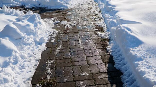 Winter sidewalk covered in icy potholes. Ice and white snow on the uncleared asphalt. Snowdrifts to the left and right of narrow road. A sunny, frosty January day. Risk of fractures. Urban economy