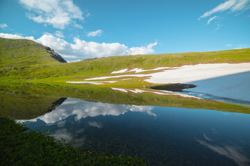 Tranquil scenery with puffy clouds in blue sky, flowering meadow, green hills and white snows reflected in calm alpine lake in sunny day. Colorful view to mountain lake and grassy hills in bright sun.
