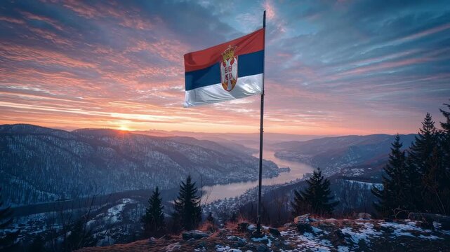 A flag of Serbia stands on a pole above a hilly landscape with winding rivers below. The view includes a small town set against rolling hills and mountains under a cloudy sky.