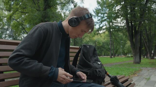 young white man on bench unpacking audio gear and cables from backpack, connecting microphone and tripod, wearing headphones, focused setup for field recording in green park with overcast sky,