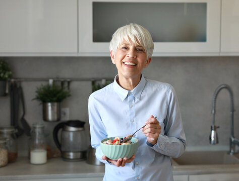 Portrait of a mid aged mature woman gettyng ready for work eating breakfast oatmeal in a bowl standing in kitchen home. Overworked, busy, stress, urgency, multitasking, active healthy senior  concepts