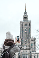 Winter city wanderlust: female tourist walking historic Warsaw old town, pastel facades and holiday lights, cozy coat, scarf and beanie. Urban sightseeing, slow travel, sustainable Europe escape eco.