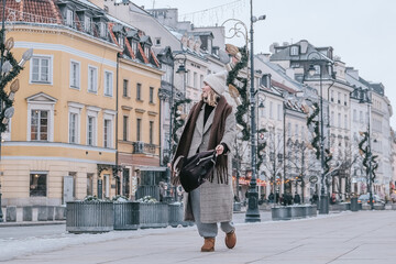 Winter city wanderlust: female tourist walking historic Warsaw old town, pastel facades and holiday lights, cozy coat, scarf and beanie. Urban sightseeing, slow travel, sustainable Europe escape eco.