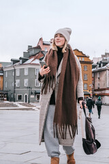 Winter city wanderlust: female tourist walking historic Warsaw old town, pastel facades and holiday lights, cozy coat, scarf and beanie. Urban sightseeing, slow travel, sustainable Europe escape eco.