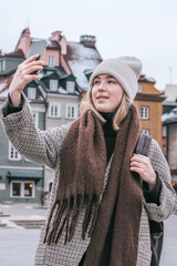 Winter city wanderlust: female tourist walking historic Warsaw old town, pastel facades and holiday lights, cozy coat, scarf and beanie. Urban sightseeing, slow travel, sustainable Europe escape eco.