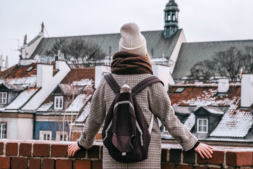 Winter city wanderlust: female tourist walking historic Warsaw old town, pastel facades and holiday lights, cozy coat, scarf and beanie. Urban sightseeing, slow travel, sustainable Europe escape eco.